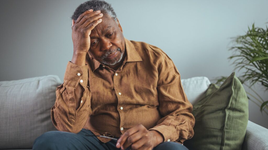 Older man sitting on a couch with his hand on his forehead, showing stress linked to retirement loneliness.