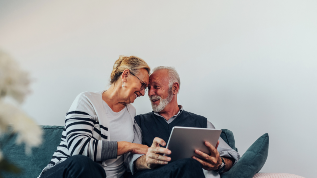 Happy retired couple laughing together on a couch while reviewing retirement plans on a tablet